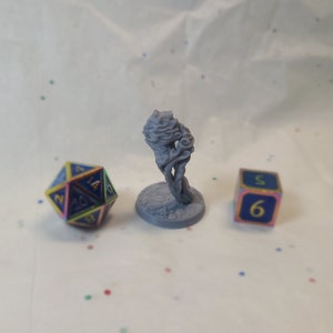 Könnte beinhalten: Eine graue 3D-gedruckte Miniatur eines Wesens mit einem wirbelnden, strukturierten Design. Die Miniatur steht auf einem runden Sockel und befindet sich neben zwei blauen Würfeln mit gelben Zahlen. Die Würfel zeigen die Zahlen 2 und 6.