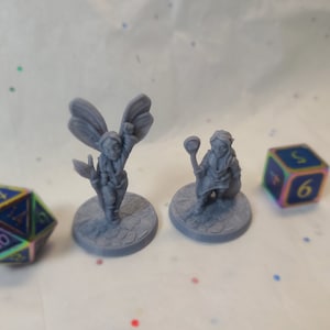 Op de afbeelding: Twee grijze 3D-geprinte miniatuurfiguurtjes op ronde bases. Het figuurtje links heeft vleugels en houdt een staf vast. Het figuurtje rechts houdt een staf vast en draagt een gewaad. Een veelkleurige 20-zijdige dobbelsteen bevindt zich op de voorgrond.