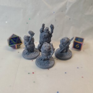 May include: Four gray 3D printed miniature figurines of frogs wearing different outfits and holding various objects. The figurines are standing on round bases. There are two dice in the background.