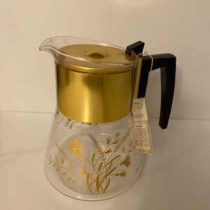 Peut inclure: Une cafetière en verre transparente et dorée avec une poignée noire et un motif floral doré. La cafetière a un couvercle doré et une étiquette attachée à la poignée.