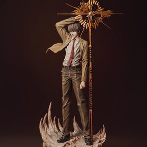 May include: A detailed anime-style figurine of a person with brown hair wearing a tan blazer, white dress shirt, and red tie. The figure stands on a textured base with skull-like details, holding a tall, ornate gold staff with a starburst top.