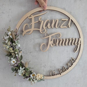Könnte beinhalten: Ein hölzernes, kreisförmiges Hochzeitsschild mit den Namen "Franzi & Tommy" und dem Datum "05. Juli 2024" ausgeschnitten. Das Schild ist mit einem Blumengesteck aus getrockneten weißen, grünen und lila Blumen und Blättern verziert.