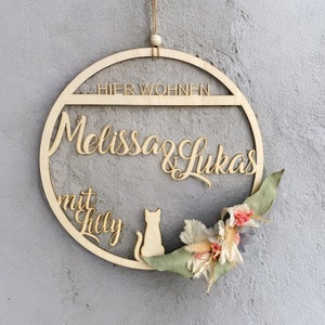 Könnte beinhalten: Ein rundes Holzschild mit dem Text "Hier Wohnen Melissa & Lukas mit Lilly" und einer Katzensilhouette. Das Schild ist mit getrockneten Blumen und Grünpflanzen verziert.