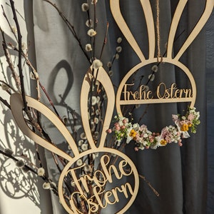 Könnte beinhalten: Zwei hölzerne Osterdekorationen in Hasenform mit dem Text "Frohe Ostern" in einem Kreis. Die Dekorationen hängen an einem Zweig mit weißen, flauschigen Blüten.