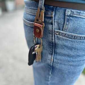 May include: A bronze metal key clip with a brown leather detail is attached to a belt loop on blue denim jeans. A set of keys and a black car key fob are suspended from the clip.