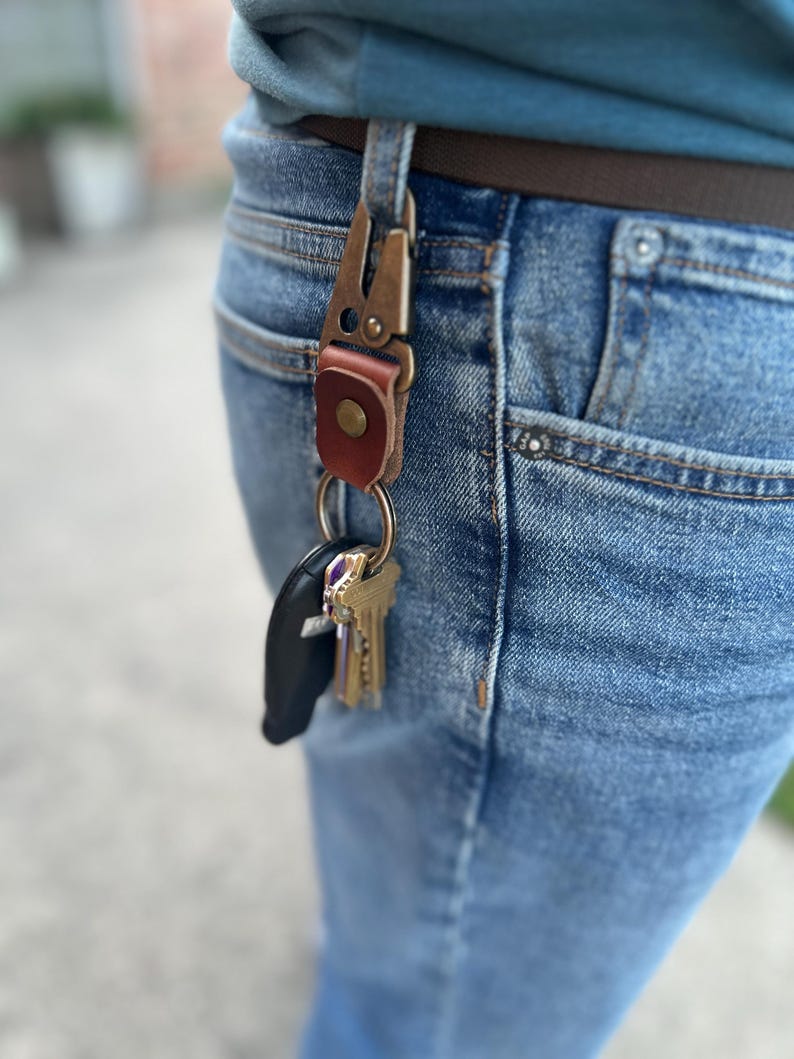 May include: A bronze metal key clip with a brown leather detail is attached to a belt loop on blue denim jeans. A set of keys and a black car key fob are suspended from the clip.