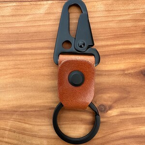 May include: A black metal carabiner with a brown leather strap and a black split ring. The carabiner has a spring-loaded gate and a round hole. The leather strap is secured with a black press stud.