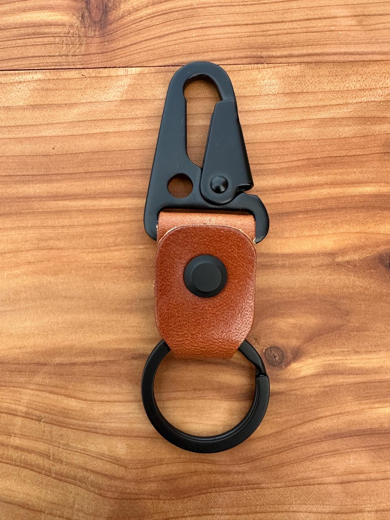 May include: A black metal carabiner with a brown leather strap and a black split ring. The carabiner has a spring-loaded gate and a round hole. The leather strap is secured with a black press stud.