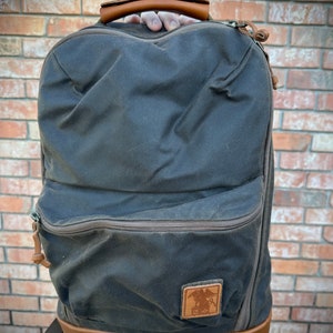 May include: A dark brown backpack with a leather handle and base. The backpack has a front pocket and a leather patch with a logo. The zippers and trim are brown. The backpack is made of a durable, water-resistant material.