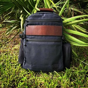 May include: A navy blue backpack with brown leather accents. The backpack features multiple zippered compartments, a brown leather patch, and a top handle. It is set against a backdrop of green foliage and grass.