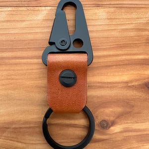 May include: A black metal carabiner keychain with a brown leather strap and a black key ring. The carabiner has a spring-loaded gate and a swivel base. The leather strap is secured with a black screw.
