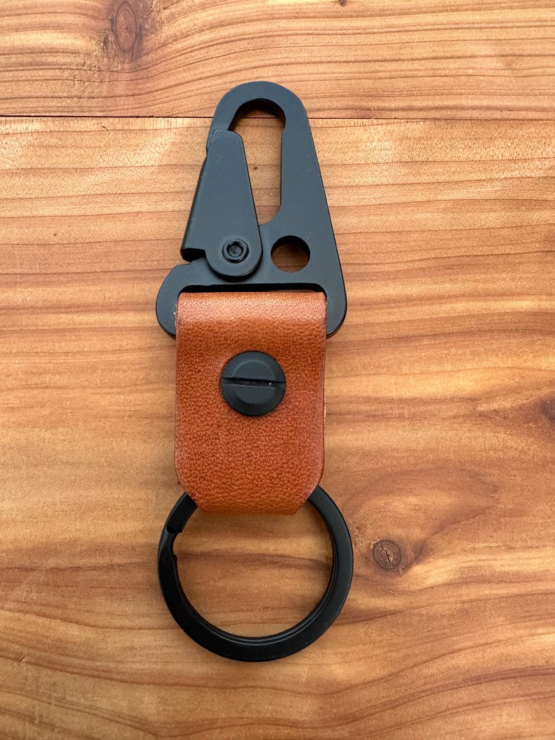 May include: A black metal carabiner keychain with a brown leather strap and a black key ring. The carabiner has a spring-loaded gate and a swivel base. The leather strap is secured with a black screw.