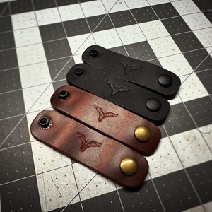 May include: Four leather cable organizers in black and brown. Each organizer features a snap closure and an embossed bird design. The brown organizers have brass-colored snaps, while the black ones have black snaps.