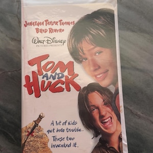 May include: A VHS tape for the Disney movie "Tom and Huck" featuring Jonathan Taylor Thomas and Brad Renfro. The cover has images of the actors and the film's title in red. The text "A lot of kids get into trouble. These two invented it." is also visible.