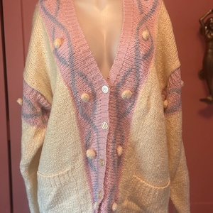 May include: A vintage, button-down cardigan in cream, pink, and gray. The sweater features a V-neck, two front pockets, and decorative pom-poms. The design includes pink and gray patterned accents on the front and sleeves. The cardigan is displayed on a mannequin.