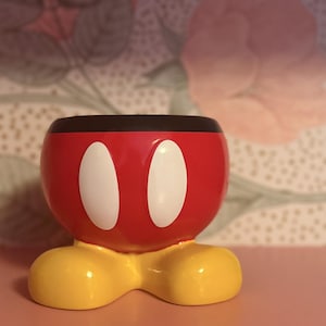 May include: A red and black Mickey Mouse-themed planter with two white oval shapes and yellow feet. The planter is a decorative item, likely for small plants or storage. The design is a playful nod to the iconic Disney character.