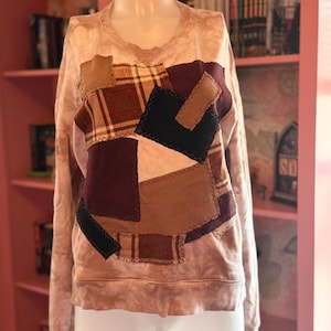 May include: A light brown sweatshirt with a patchwork design. The patchwork includes squares of plaid, corduroy, and solid fabrics in shades of brown, burgundy, and black. The sweatshirt has a crew neck and long sleeves.