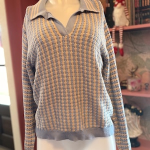May include: A light blue and tan knit sweater with a V-neck collar and long sleeves. The sweater has a textured pattern and is displayed on a mannequin. The collar and cuffs are a solid light blue.