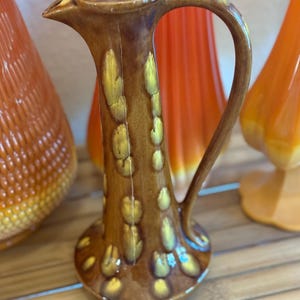 May include: A brown ceramic pitcher with a handle and a spout. The pitcher has a textured surface with yellow accents. Other orange and yellow vases are in the background.