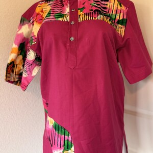 May include: A magenta short-sleeved shirt with a mandarin collar and a button-down placket. The shoulders and side panels feature a colorful, abstract floral print with pink, yellow, green, and black accents. The shirt has a relaxed fit.