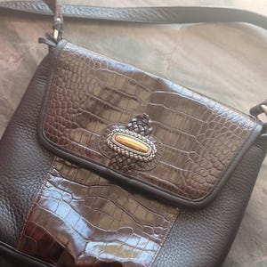 May include: A brown leather crossbody bag with a faux crocodile skin flap and centre panel. The bag features a decorative silver and gold clasp and a long, adjustable strap. The bag is made of textured leather.