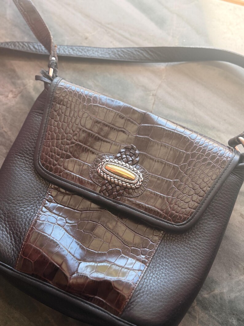 May include: A brown leather crossbody bag with a faux crocodile skin flap and centre panel. The bag features a decorative silver and gold clasp and a long, adjustable strap. The bag is made of textured leather.