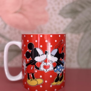 May include: Red ceramic mug with white polka dots featuring Mickey and Minnie Mouse kissing. Mickey is wearing red shorts and yellow shoes, while Minnie wears a blue polka dot skirt and a red bow. The characters are forming a heart with their hands.