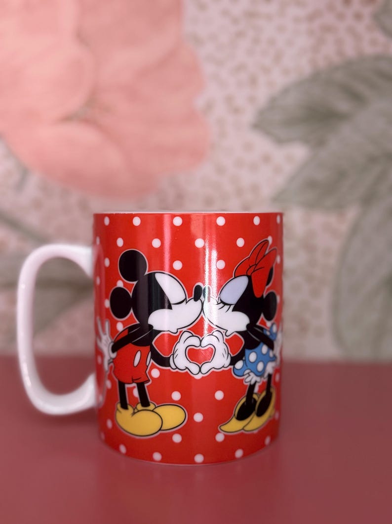 May include: Red ceramic mug with white polka dots featuring Mickey and Minnie Mouse kissing. Mickey is wearing red shorts and yellow shoes, while Minnie wears a blue polka dot skirt and a red bow. The characters are forming a heart with their hands.