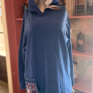May include: A navy blue hoodie with a cream-colored hood lining. The hoodie features a pocket with nautical-themed patches, including an anchor, stars, and a sailboat. The long-sleeved top is displayed on a mannequin.