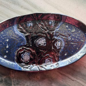 May include: An oval-shaped ceramic serving dish with a glossy finish. The dish features a deep blue interior with white speckles and a raised, textured floral design in the center. The rim is a metallic silver color, and the exterior is a deep red.