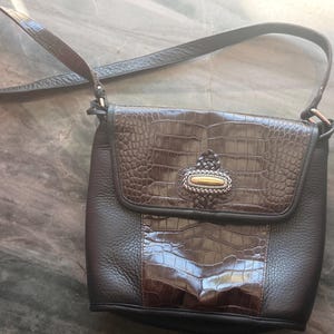 May include: A brown leather crossbody bag with a textured flap and a decorative clasp. The bag features a crocodile-like embossed pattern on the flap and a contrasting smooth leather body. The adjustable strap is also brown.