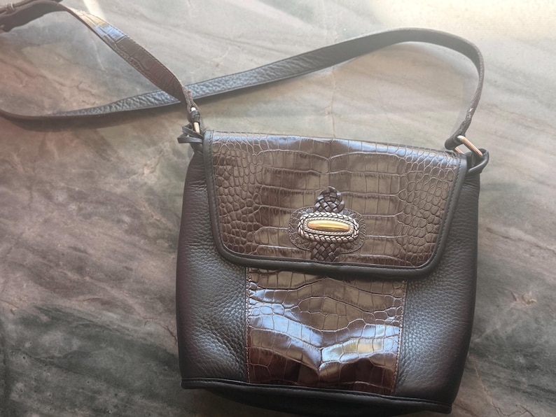 May include: A brown leather crossbody bag with a textured flap and a decorative clasp. The bag features a crocodile-like embossed pattern on the flap and a contrasting smooth leather body. The adjustable strap is also brown.