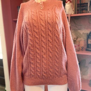 May include: A dusty rose cable-knit sweater with a mock neck and three pearl buttons. The sweater has long sleeves and a ribbed hem and cuffs. The sweater is displayed on a mannequin.