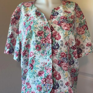 May include: A short-sleeved blouse with a floral pattern. The fabric is white with a vibrant print of pink, red, purple, and blue flowers, along with green leaves. The blouse has a V-neck and button closure.