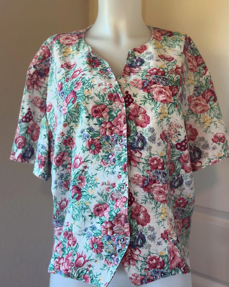 May include: A short-sleeved blouse with a floral pattern. The fabric is white with a vibrant print of pink, red, purple, and blue flowers, along with green leaves. The blouse has a V-neck and button closure.