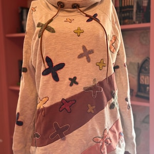 May include: A light gray sweatshirt with a cowl neck and brown drawstrings. The sweatshirt features colorful cross-shaped appliqués in various colors, including blue, green, and pink, and a diagonal stripe of maroon fabric.
