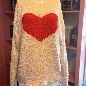 May include: Cream-colored sweater with a large red heart design on the front. The sweater has a textured, fuzzy appearance and long sleeves. The heart is a solid, vibrant red color.