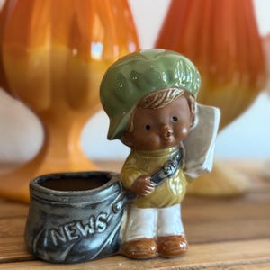 May include: A vintage ceramic figurine of a newsboy, wearing a green cap, yellow shirt, and white pants. The newsboy holds a newspaper and a bag with the word "NEWS" on it. The figurine is a planter, with a small opening for plants.