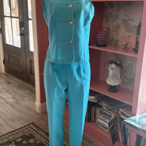 May include: A turquoise two-piece suit displayed on a mannequin. The suit includes a collared, sleeveless vest with gold buttons and matching straight-leg pants. The outfit is made of a smooth, solid-colored fabric, suggesting a vintage or retro style.