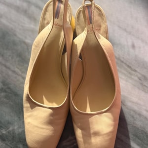May include: A pair of light beige slingback heels with square toes. The shoes have a strap around the heel and a small heel. The interior of the shoe is a lighter shade of beige. The brand name is visible on the insole.
