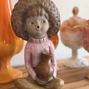 May include: A ceramic figurine of a seated figure wearing a large, textured hat and a pink sweater. The figure holds a brown jug and is seated with legs crossed. The figurine has a neutral skin tone and is on a wooden surface.