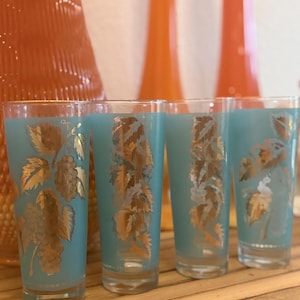 May include: Four vintage turquoise highball glasses with gold leaf and grape vine designs. The glasses are arranged on a wooden surface, with orange decorative items in the background. The glasses are approximately 6 inches tall.