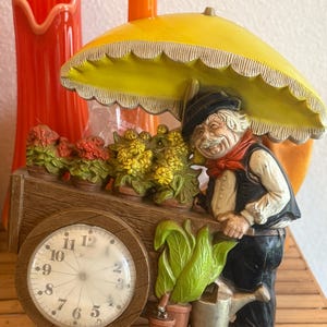 May include: A decorative clock featuring a figurine of a flower vendor pushing a cart filled with potted flowers. The vendor is under a yellow umbrella. The clock face is white with black numerals and hands.