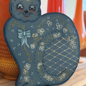 May include: A hand-painted, blue cat-shaped wooden decoration. The cat features a bow, floral accents, and a lattice design on its tail. The cat's face has rosy cheeks and a smiling expression. The decoration is displayed on a wooden surface.