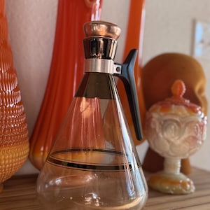 May include: A vintage glass carafe with a copper-colored lid and a black handle. The carafe has a clear, conical body with a black and gold band. Other orange glass objects are in the background, creating a retro aesthetic.