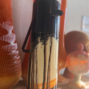 May include: A vintage black and white thermos with a landscape design featuring trees and a sunset. The thermos has a black handle and a push-button top. The background includes orange vases.