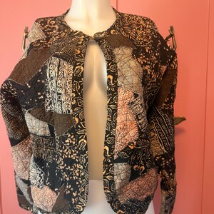 May include: A cropped jacket featuring a patchwork design with various black, brown, and tan fabric patterns. The garment has long sleeves, a round neckline, and a single button closure at the top. The fabric displays a mix of geometric, floral, and abstract prints throughout the quilted construction.