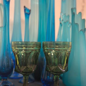 May include: Two green-tinted glass goblets with a diamond pattern are in the foreground. Behind them are various tall, slender, and decorative blue glass vases. The vases vary in shape and shade of blue, creating a visually appealing display.