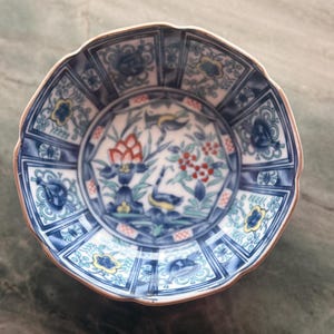 May include: A small, antique ceramic plate with a scalloped edge. The plate features a detailed, hand-painted design with blue, white, yellow, and red floral and bird motifs. The rim has a brown border.