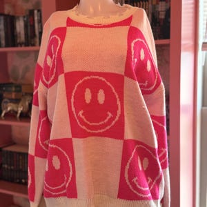 May include: A cream-colored sweater with a pink checkerboard pattern featuring smiley faces. The sweater has a distressed neckline and hem. The long-sleeved knitwear is displayed on a mannequin, showcasing its design and texture.